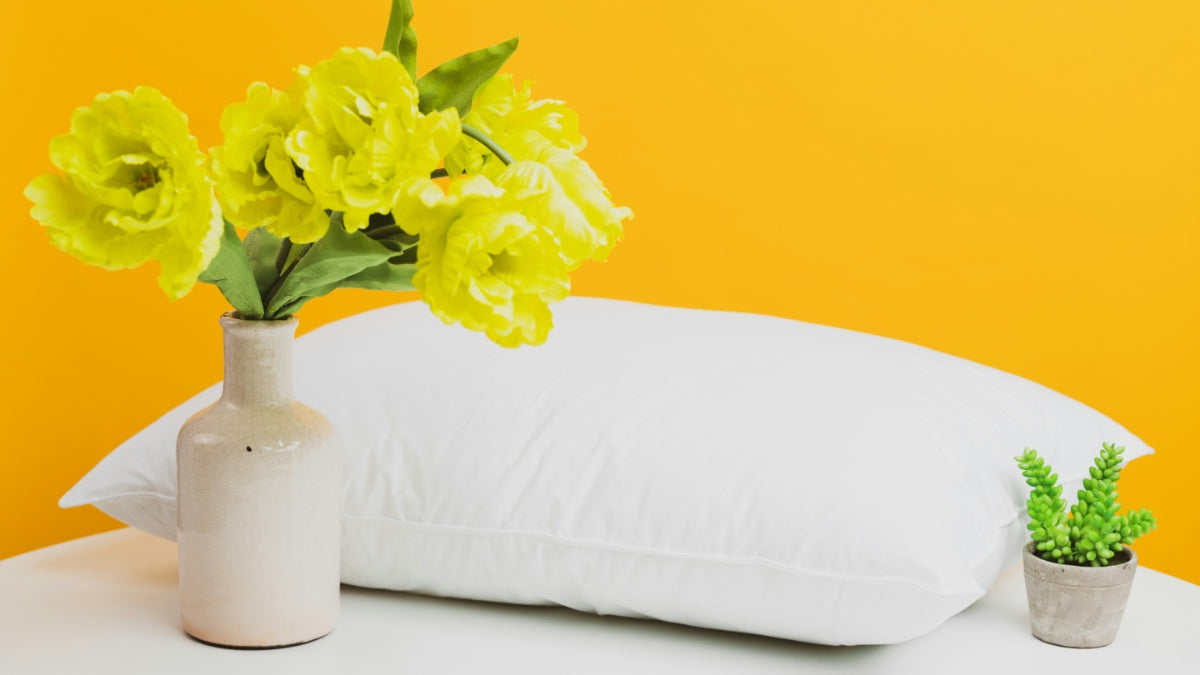 A white pillow against a yellow background next to a vase of flowers and plant pot