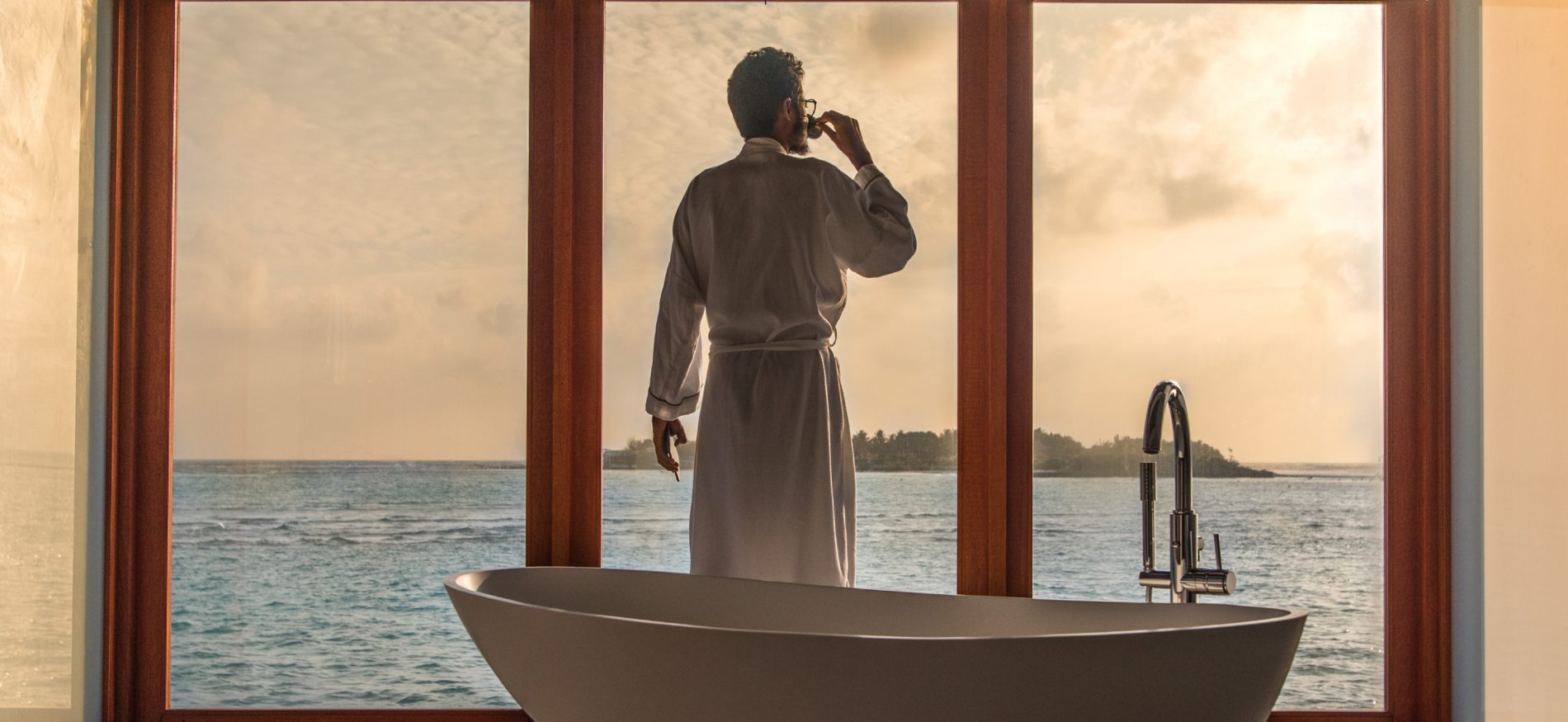 Man looking out to sunset, dressed in a luxurious bathrobe and beside a spectacular bath, after getting up from bed to have a cup of tea or coffee.