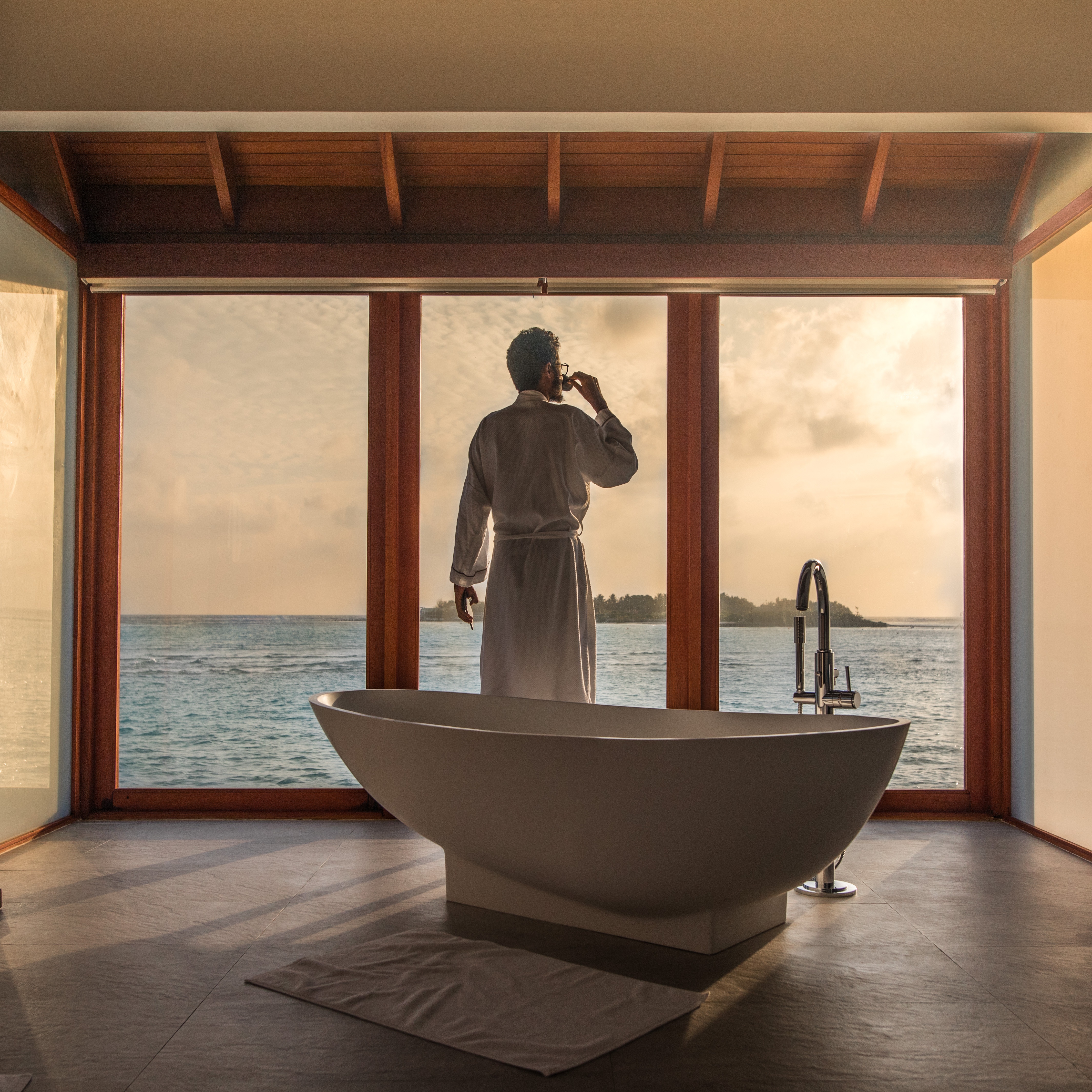 Man looking out to sunset, dressed in a luxurious bathrobe and beside a spectacular bath, after getting up from bed to have a cup of tea or coffee.