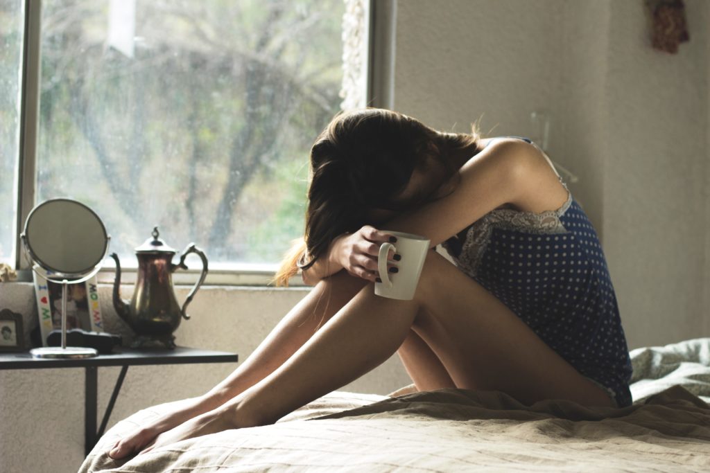 Girl in bed, upset whilst holding cup of tea or coffee. Dr. Alanna Hare recommends scheduling "worry time" each evening, to prevent rumination before bedtime.