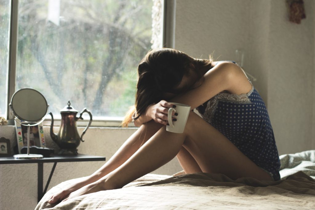 Girl in bed, upset whilst holding cup of tea or coffee. Dr. Alanna Hare recommends scheduling "worry time" each evening, to prevent rumination before bedtime.